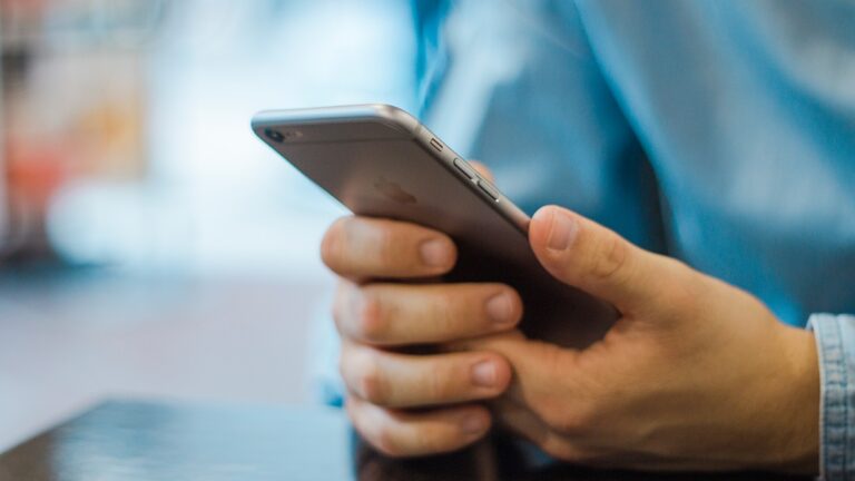 A person in a blue shirt, inspired by Kyles Story, is holding and using a silver smartphone with both hands while sitting at a dark-colored table. The background is blurred.