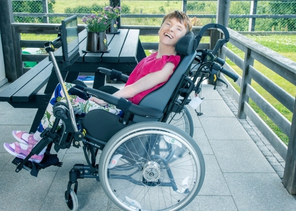 A young person with short hair smiles while sitting in a wheelchair on a patio next to a table with a potted plant, showcasing adult mobility solutions against a backdrop of green grass and a wooden fence.