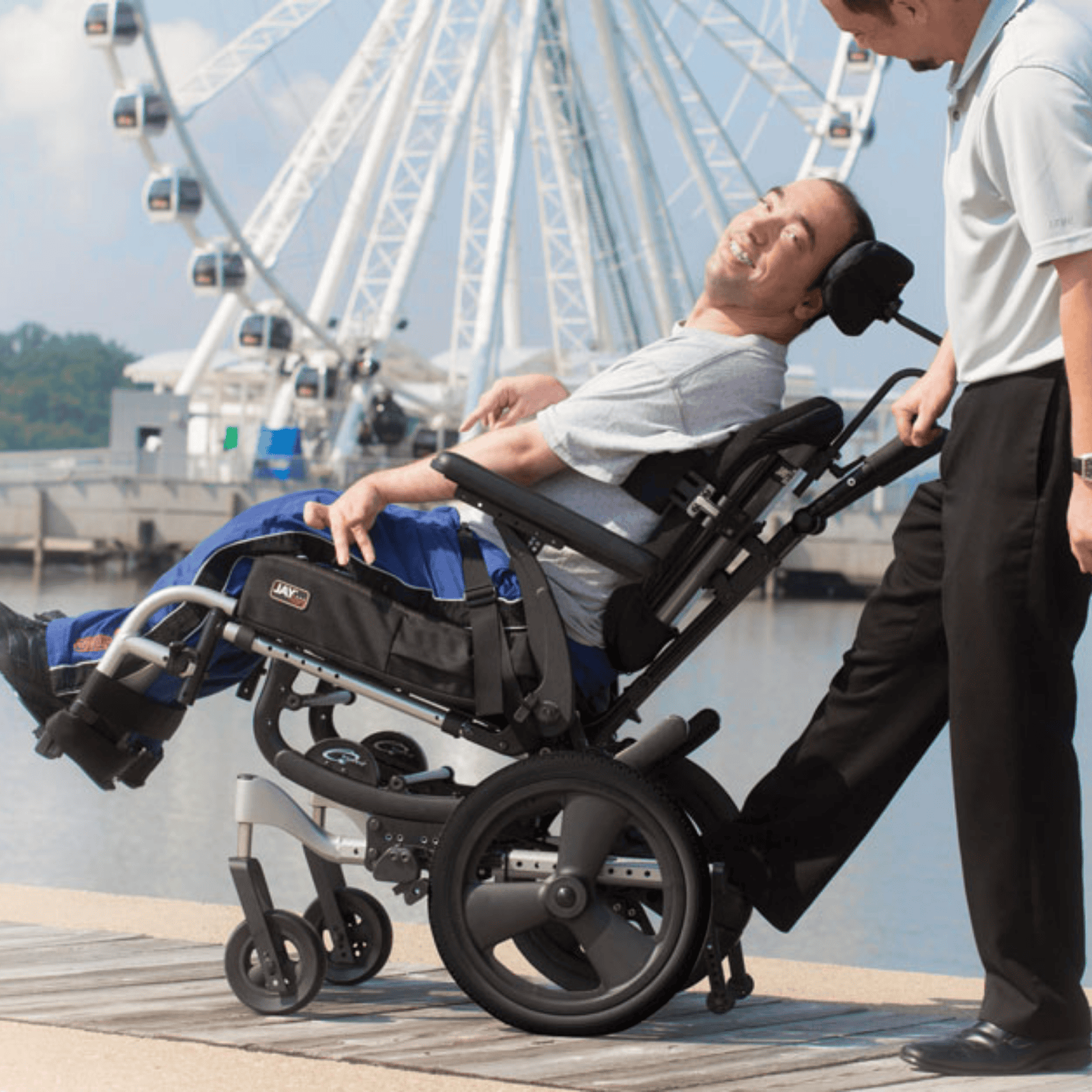 A man reclines in a tilt-in-space wheelchair on a dock, smiling at a standing caregiver. A large Ferris wheel is visible in the background near the water.