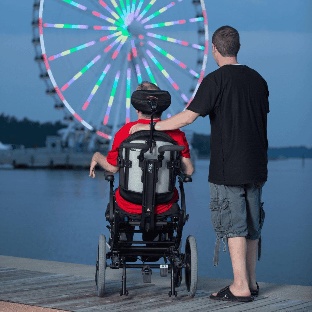 A man stands beside a person using a tilt-in-space wheelchair, both facing a waterfront at dusk. In the background, a large Ferris wheel glows with colorful lights against the evening sky.