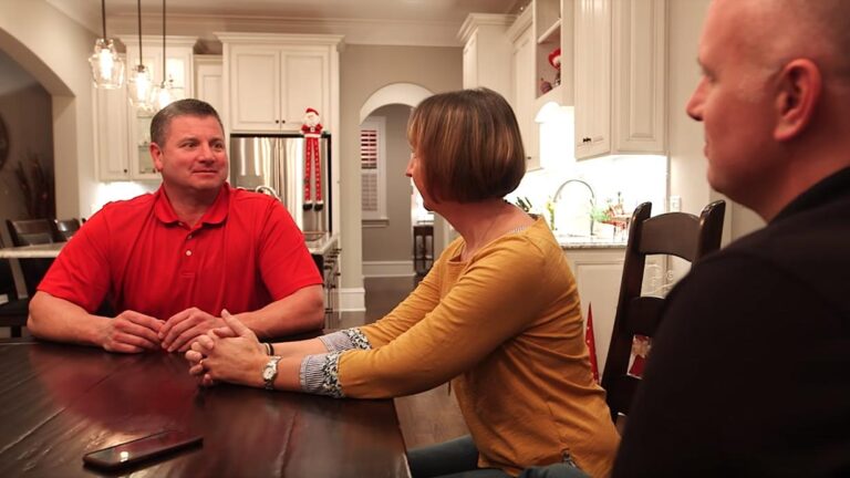 Three adults sit around a wooden table in a kitchen, engaged in conversation. The man on the left wears a red shirt, the woman in the center wears a yellow top, and the man on the right wears black—each sharing moments from Todds Story.