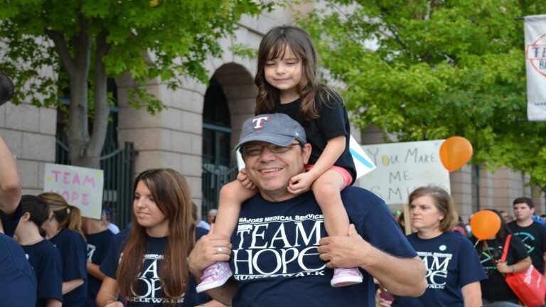 A man wearing glasses and a Team Hope shirt smiles with a young girl on his shoulders at an outdoor event, surrounded by people in matching shirts holding awareness signs and balloons. Trees and a building are in the background.