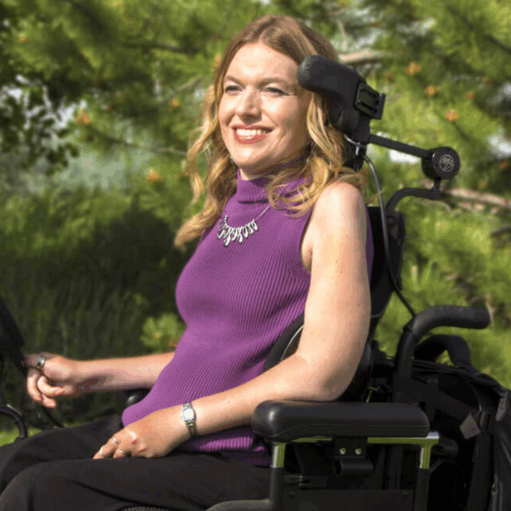 A woman with blonde hair in a purple sleeveless top sits in a motorized wheelchair with alternative access controls outdoors, smiling, surrounded by green trees and plants.