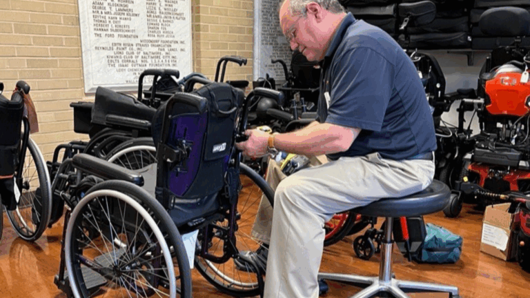 A man sits on a stool indoors, focused on service and repair of a wheelchair with tools. Several other wheelchairs and mobility devices surround him, with a wall listing names visible in the background.
