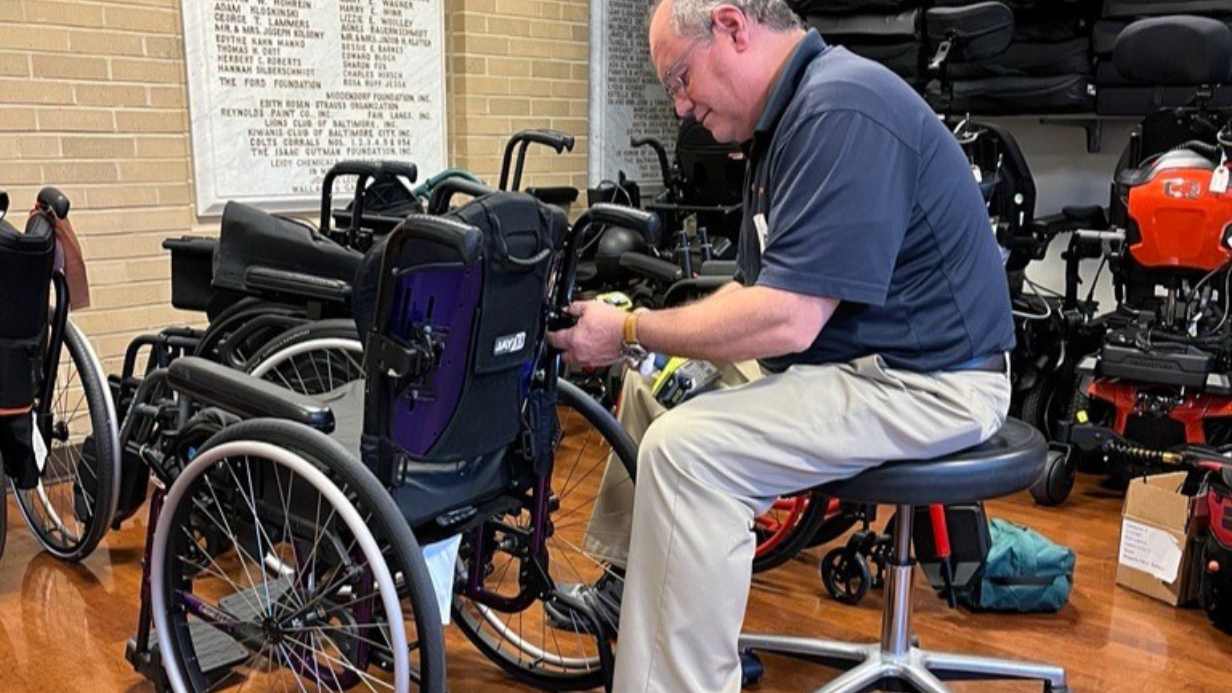 A man sits on a stool indoors, focused on service and repair of a wheelchair with tools. Several other wheelchairs and mobility devices surround him, with a wall listing names visible in the background.