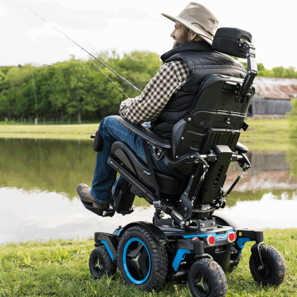A man in a plaid shirt and hat sits in a power tilt wheelchair by a pond, fishing with his rod. Green trees and a rustic barn complete the peaceful background.