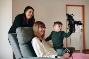 A young child stands next to an elderly woman in a chair, both looking at a tablet. A smiling woman stands behind them, watching. They appear to be enjoying a video call together with Speech Solutions in a cozy living room.