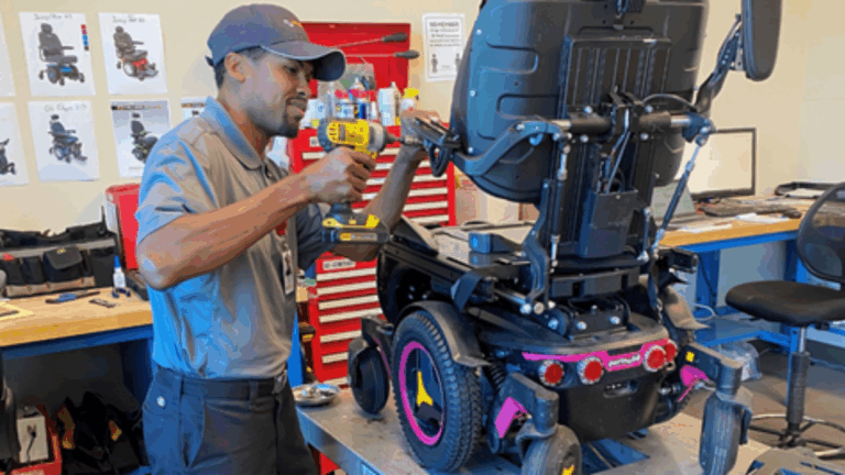 A technician in a gray uniform uses a power drill to repair an electric wheelchair in a workshop, offering in-shop service with tools and equipment visible in the background.