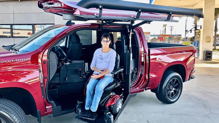 Tricias Story: A woman sits in a power wheelchair attached to the driver’s seat of a red pickup with a modified door and roof lift at a gas station. The open door reveals the impressive accessibility adaptation.