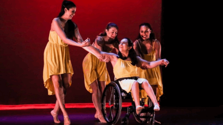 Four women in yellow dresses perform a dance on stage; one performer in Bethanys Story uses a wheelchair while the others support her, all smiling under warm lighting against a red background.