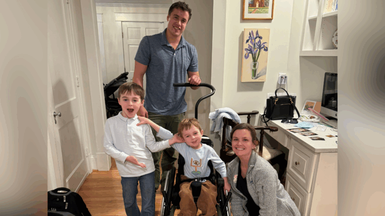 A smiling family of four poses indoors. An adult man stands behind a boy in a wheelchair, while a woman crouches beside them. Another boy stands next to the group—all appearing cheerful and relaxed in the cozy home from Blakes Story.