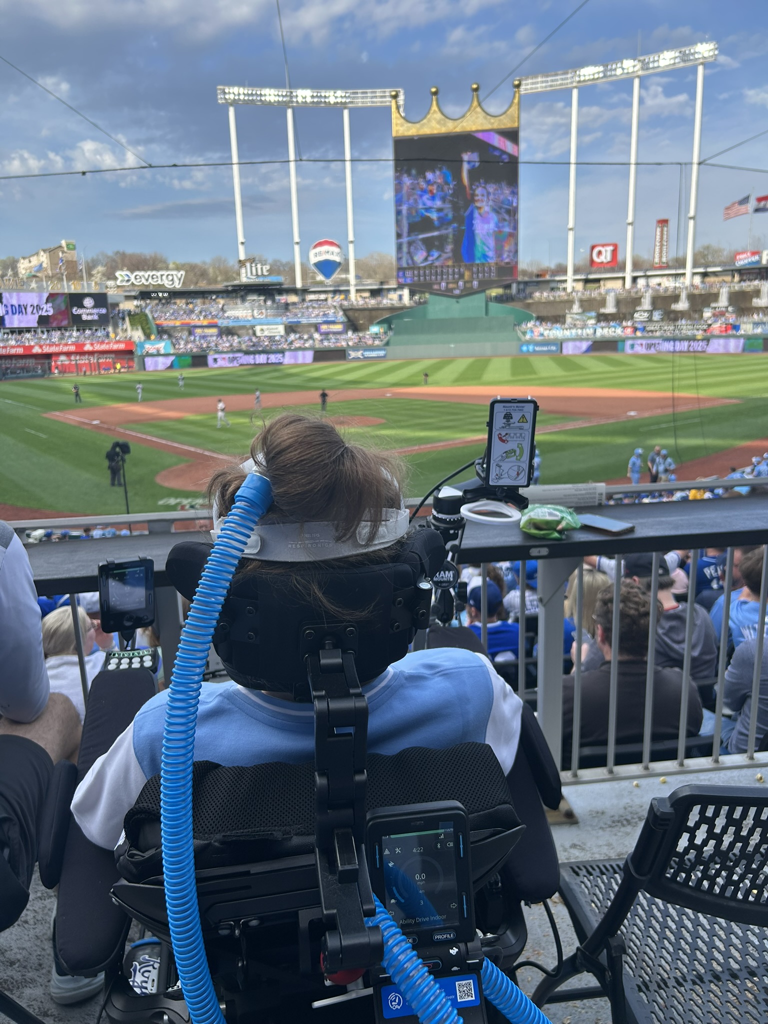 Sarahs Story unfolds as she watches a baseball game from the stands in her power wheelchair with a ventilator. The field, scoreboard, and stadium seats stretch out beneath a partly cloudy sky.