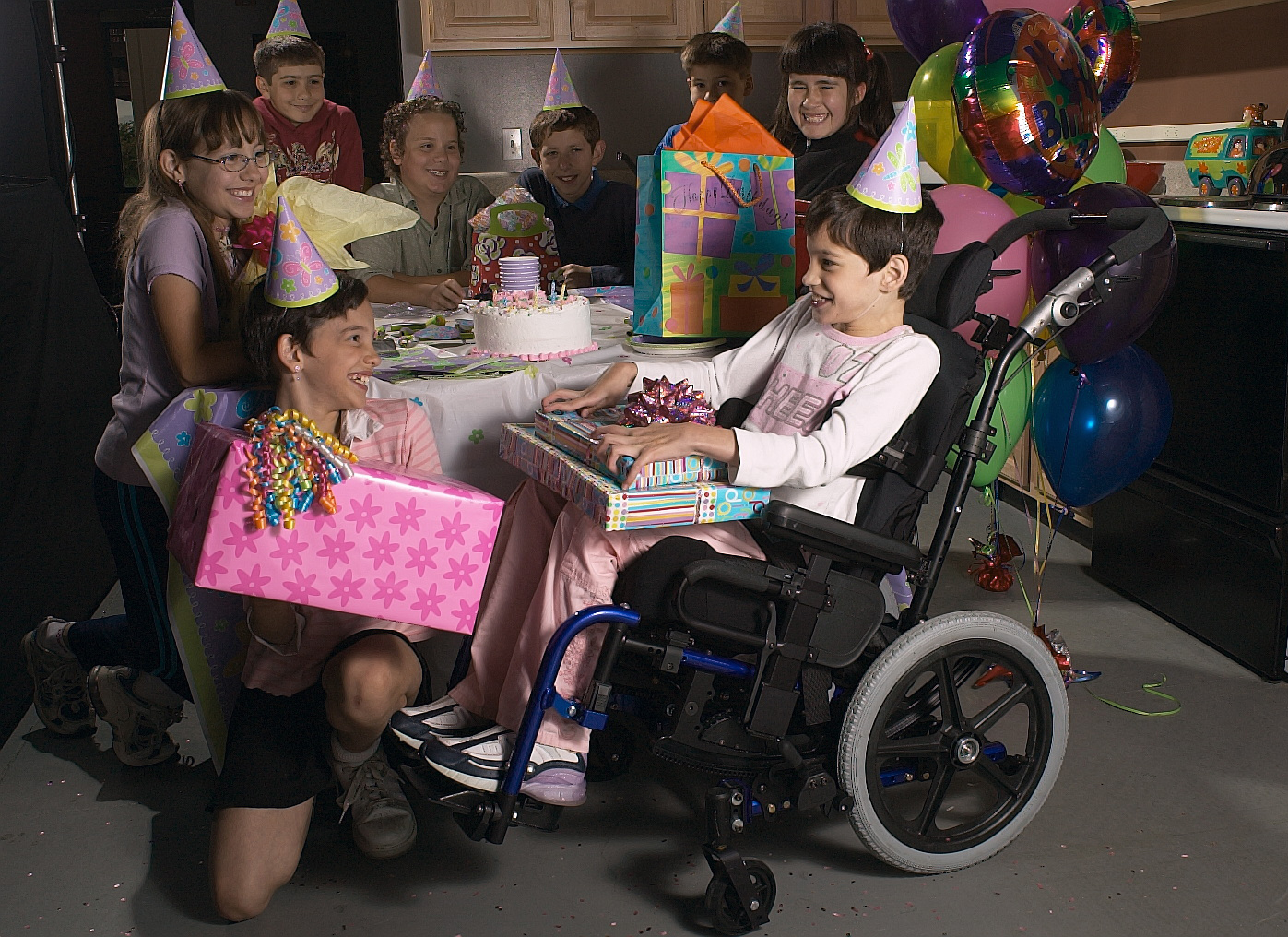 A child in a pediatric wheelchair, wearing a party hat, smiles while receiving gifts from friends at a birthday party. The group is gathered around a table with cake, balloons, and wrapped presents.