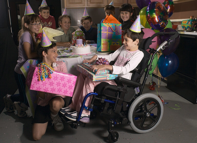 A group of children in party hats celebrate a birthday around a table with a cake. One child in a pediatric tilt wheelchair smiles and receives gifts, surrounded by friends, balloons, and colorful presents.