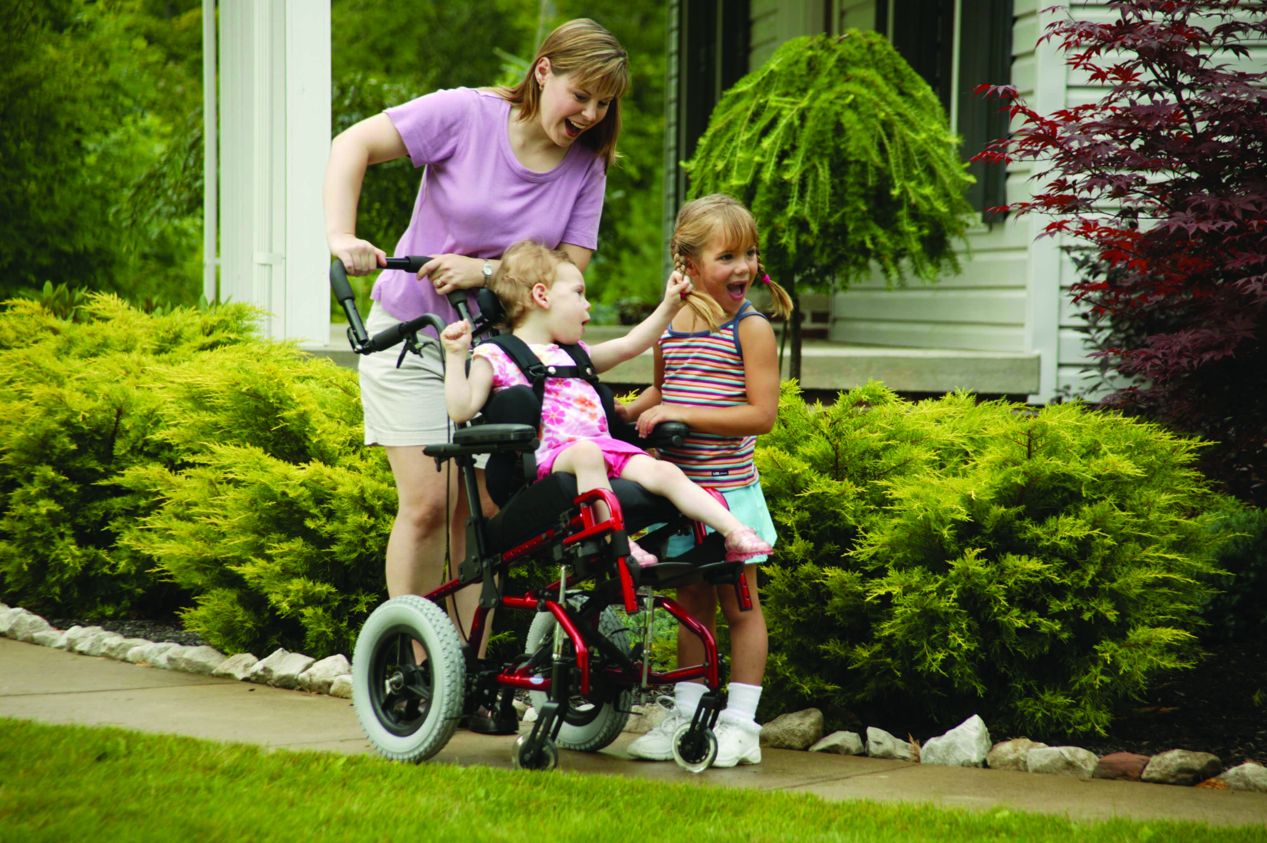 A woman pushes a young girl in a pediatric wheelchair along a path beside green bushes, while another smiling girl walks next to them, holding the wheelchair handle. All appear happy and outdoors near a house.