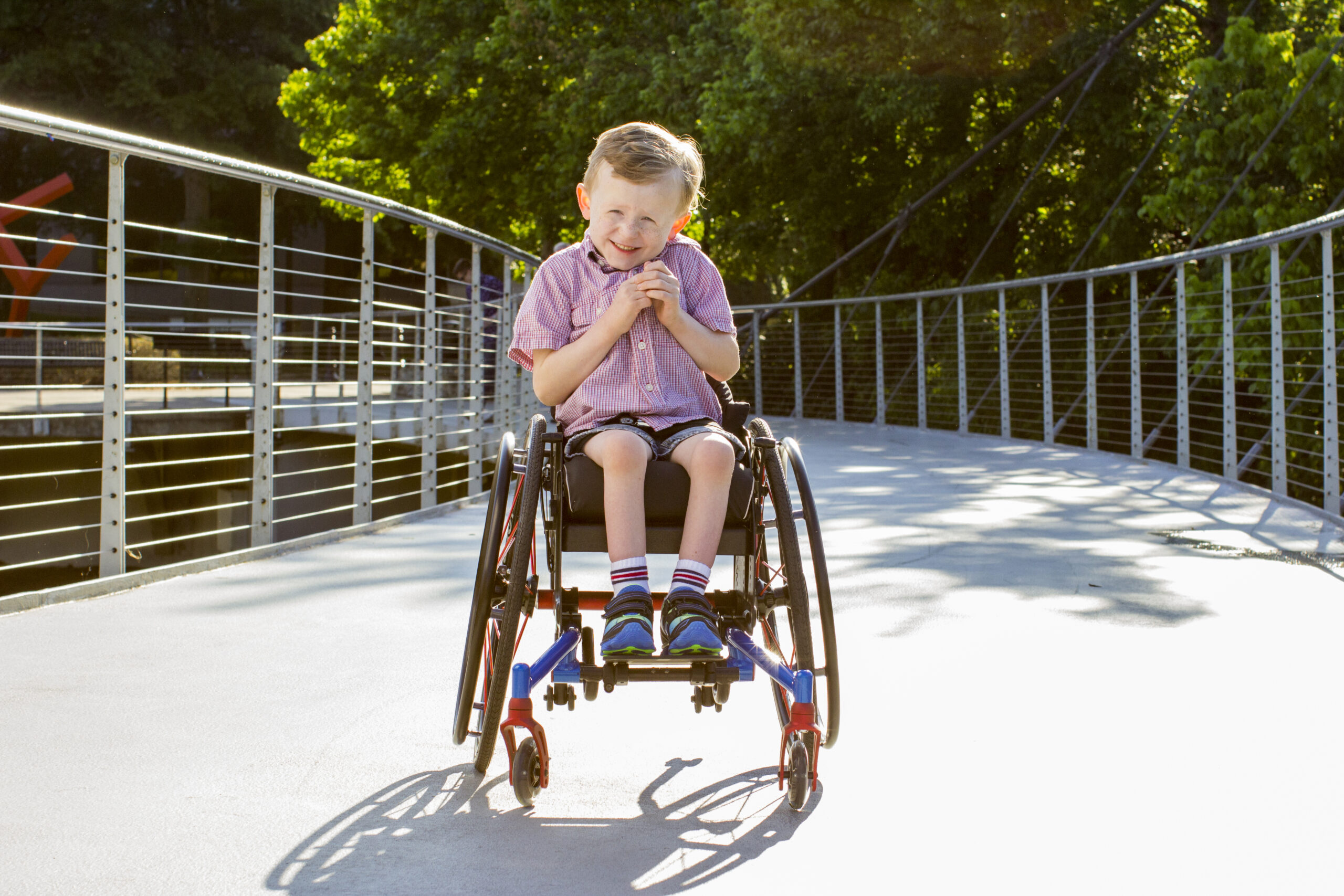 A young boy in an ultra-light wheelchair smiles and clasps his hands while sitting on a sunlit pedestrian bridge, surrounded by greenery.