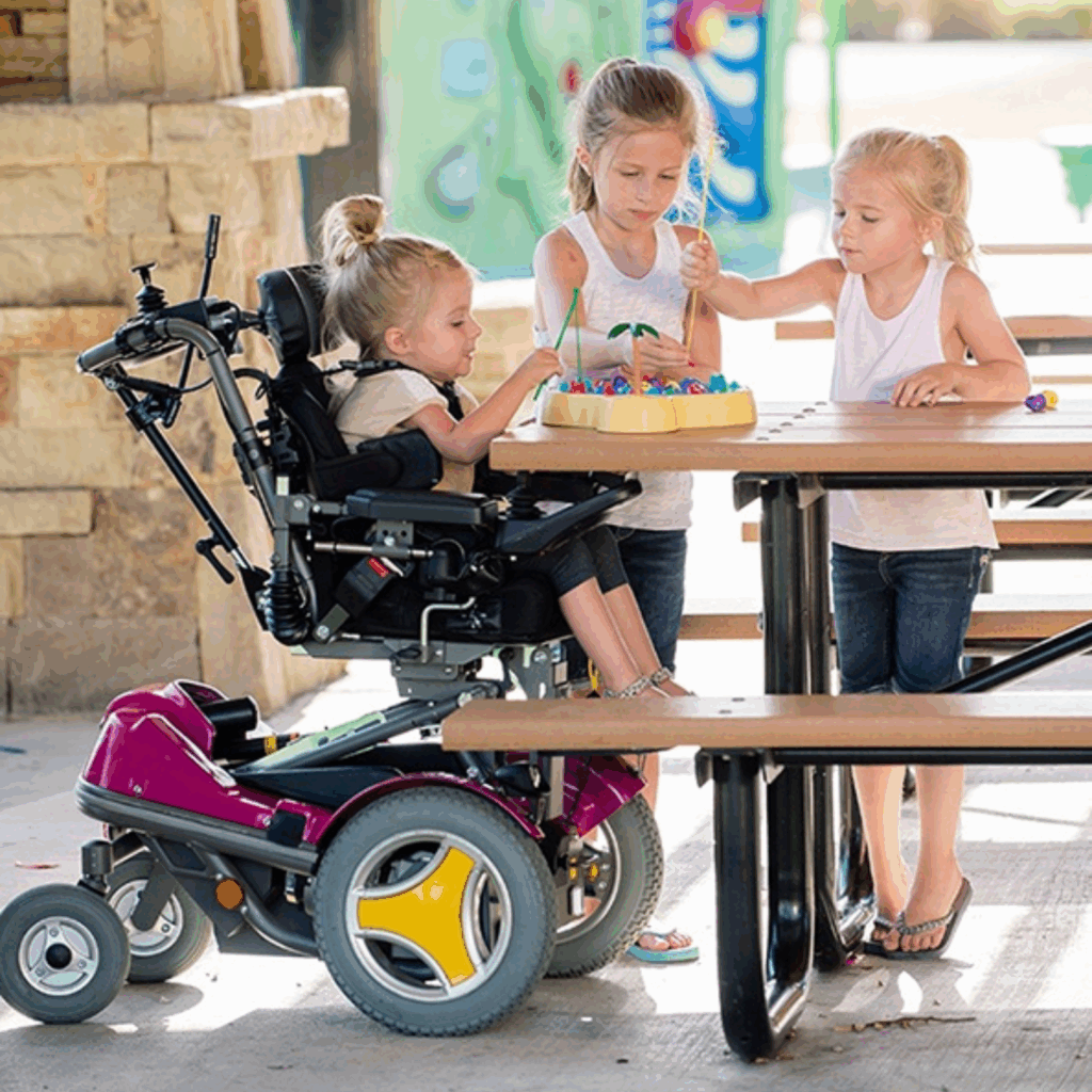 Three young girls play together at a picnic table; one girl uses a Complex Power Tilt Wheelchair, and the others stand nearby, all focused on a colorful activity in front of them.