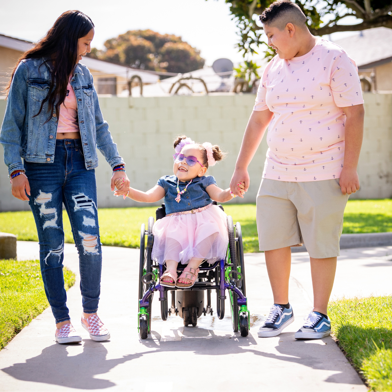 A smiling child in an ultra-light wheelchair holds hands with a woman and a boy as they walk together on a sunny sidewalk. All three look happy and casually dressed, enjoying time outdoors.