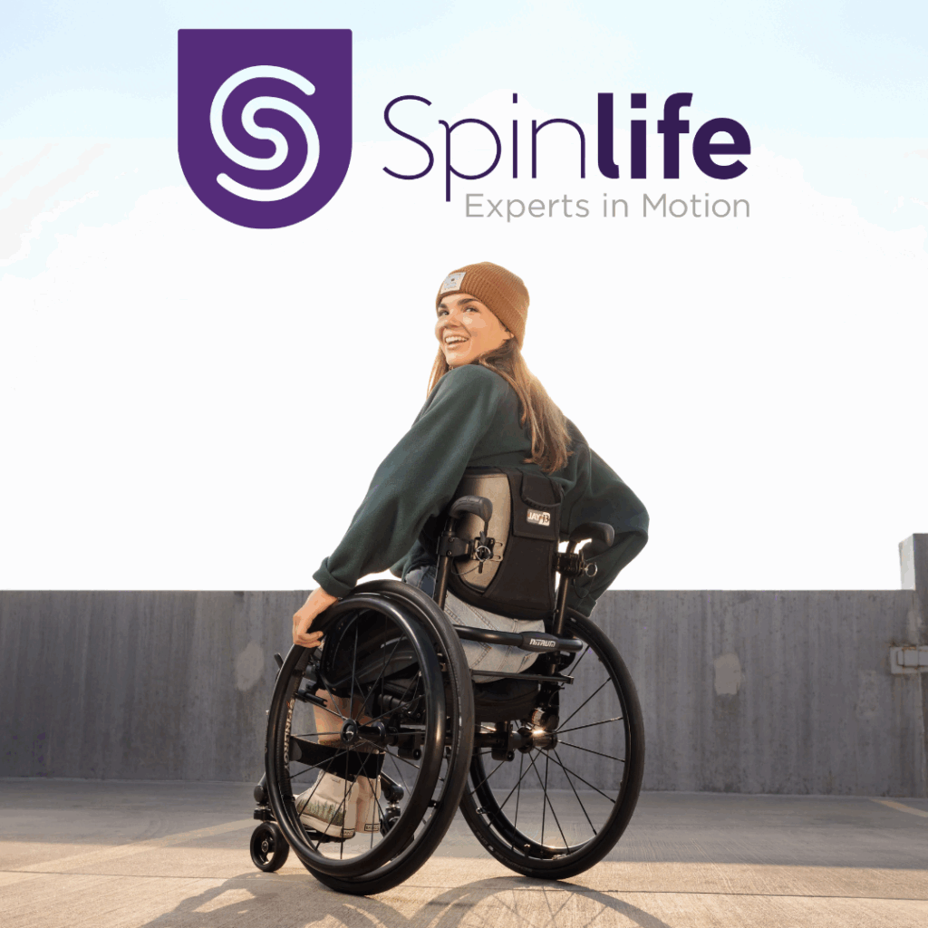 A woman using adult mobility solutions smiles and turns towards the camera outdoors, wearing a beanie and sweatshirt. Above her is the Spinlife logo with the tagline “Experts in Motion.”.