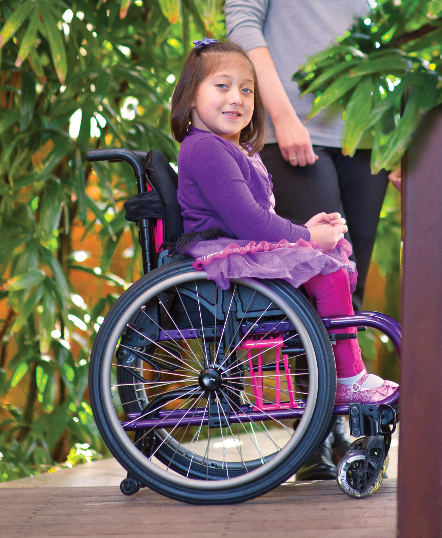 A young girl in a purple outfit and tutu smiles while sitting in a lightweight wheelchair outdoors; greenery and an adult’s legs appear in the background.