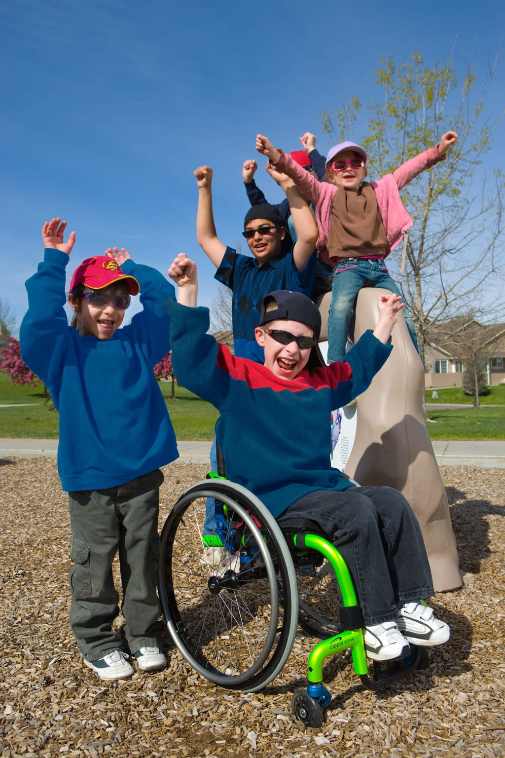 Four children, including one using an ultra-light wheelchair, smile and cheer with their hands raised while playing together at a sunny outdoor playground. Trees and houses are visible in the background.