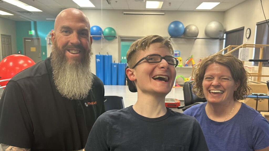 Three people smile and laugh together in a brightly lit therapy room with exercise balls and equipment in the background. The cheerful, supportive mood captures the spirit of Ryan's Story.