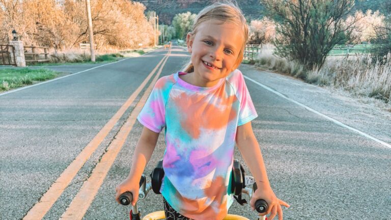 A young child in a colorful tie-dye shirt smiles while standing with a walker on a quiet country road, lined with trees and fences on a sunny day, capturing a joyful moment from Madilynns Story.