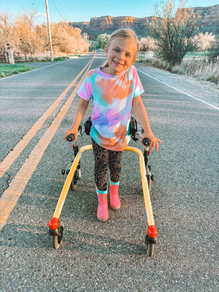 A young girl smiling, standing on a road with a yellow walker, shares Madilynns Story. She wears a colorful tie-dye shirt, patterned leggings, and pink boots. Trees and rocky hills are seen in the background under a clear sky.