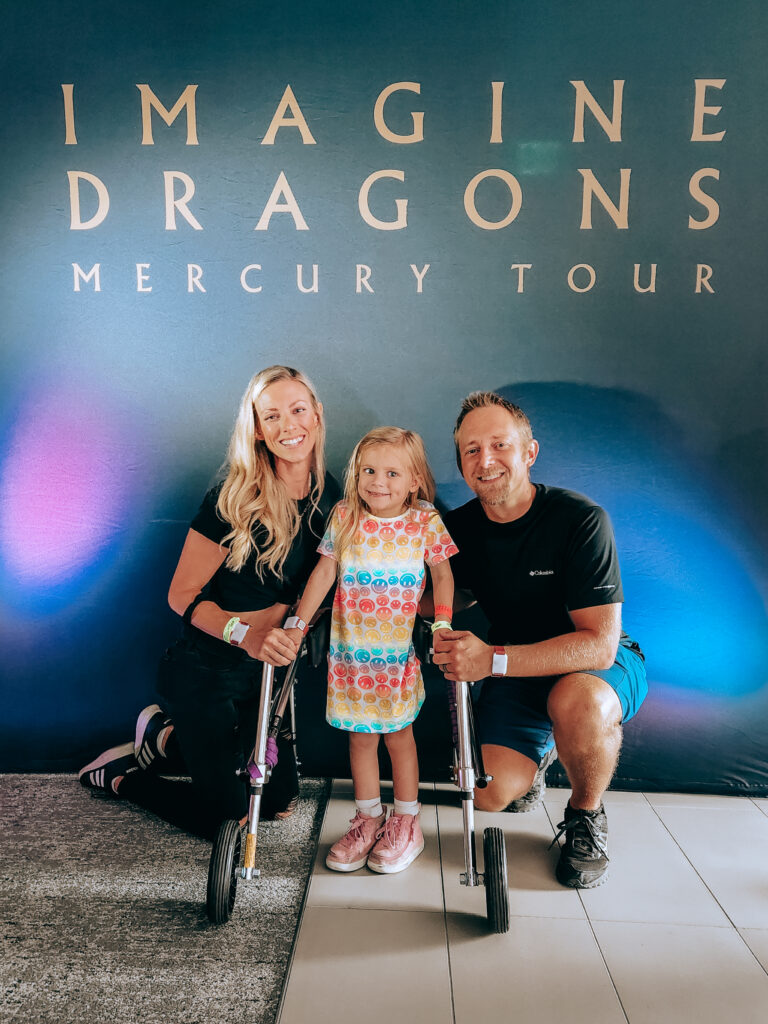 A smiling woman, young girl with a walker, and man pose together in front of a blue Imagine Dragons Mercury Tour backdrop. The cheerful and supportive atmosphere beautifully captures the spirit of Madilynns Story.