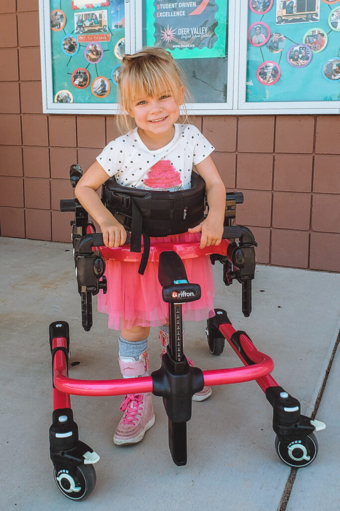 A young girl with blonde hair and bangs, wearing a white shirt, pink tutu, and pink shoes, smiles while using a pink walker outside a building with brown bricks and decorated windows—this is Madilynns Story.