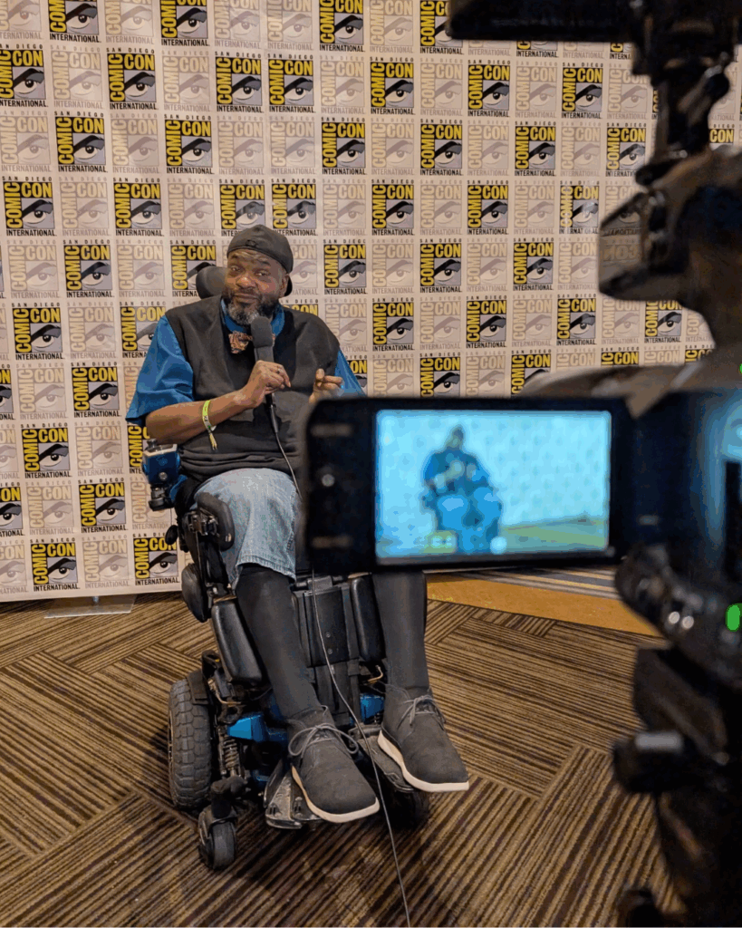 A man in a wheelchair shares Paul's Advocacy Story into a microphone before a Comic-Con backdrop, while being filmed. A camera and its screen displaying the scene are visible in the foreground.