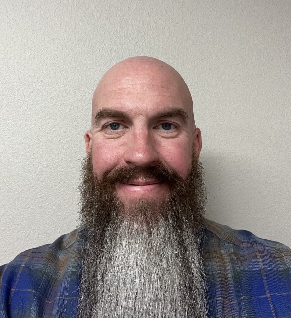 A smiling bald man with a long, full brown and gray beard, wearing a plaid shirt, stands against a plain light-colored wall—capturing a moment from Ryan's Story.