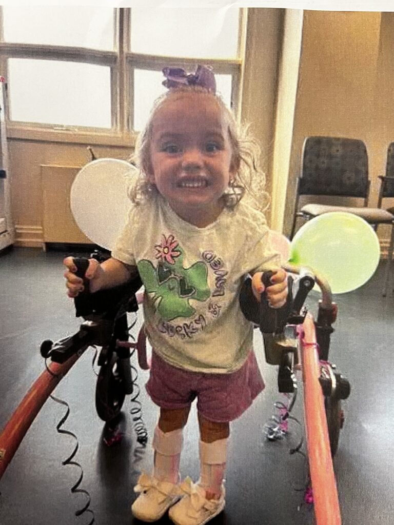 A young girl with curly hair smiles widely while using a pink walker. She wears a white shirt, pink shorts, ankle braces, and white shoes. Balloons are attached to the walker as she stands indoors, embodying the spirit of Ryan's Story.