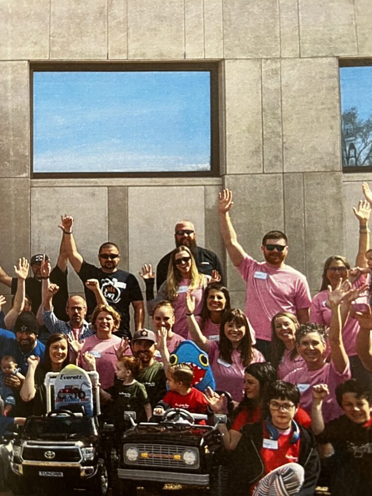 A group of people, many in pink shirts for Ryan's Story, stand and sit in front of a modern gray building, smiling and raising their hands. Two toy cars with children inside are at the front of the group.