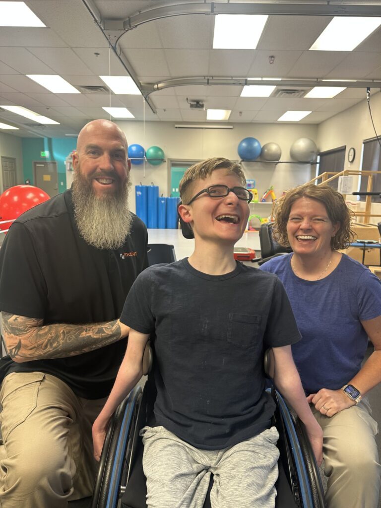 Three people are smiling in a bright physical therapy room. At the center of Ryan’s Story, a boy in a wheelchair is flanked by a bearded man and a woman. Therapy equipment and exercise balls fill the background.