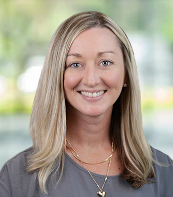 A woman with straight, shoulder-length blonde hair smiles at the camera. She is wearing a grey top, gold necklaces, and stud earrings. The softly blurred background hints at green and light—perfect for promoting comprehensive Speech Solutions coverage.