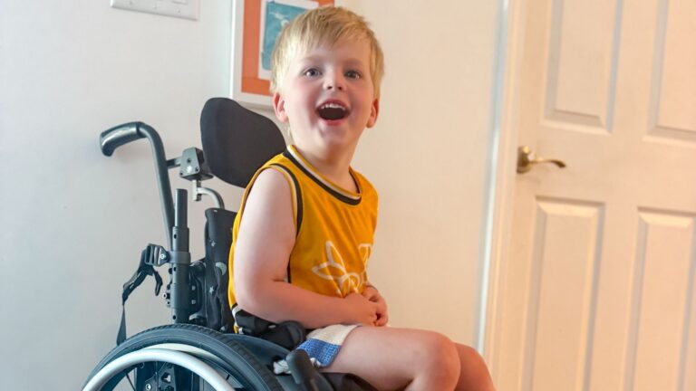 A young boy named Beckett with blond hair in a yellow tank top sits smiling in a wheelchair indoors, near a white door and a wall with framed artwork, ready to share his story.