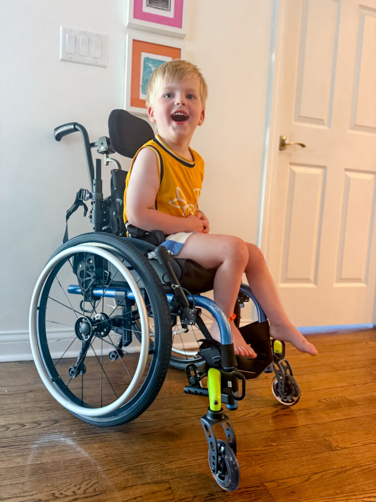 A young boy in a yellow shirt sits smiling in a wheelchair indoors, sharing Beckett's Story, with framed pictures and a closed white door in the background.