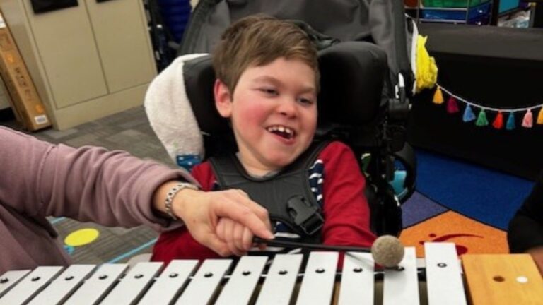 A smiling child in a wheelchair plays a xylophone with assistance from an adult, gently guiding the child’s hand with a mallet. The colorful classroom is alive with music as this becomes part of Jake’s Story.