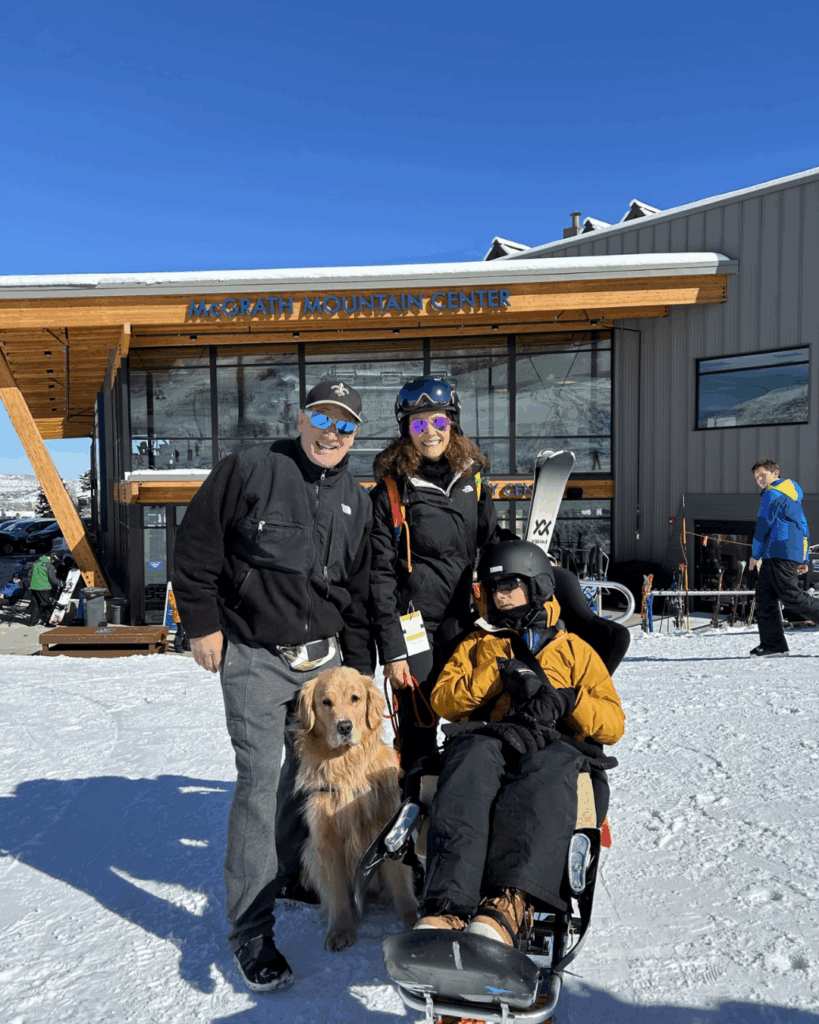 A man, a woman holding a ski, and a person in adaptive ski gear with a golden retriever pose in front of McGrath Mountain Center on a sunny, snowy day. Skis and snowboards are visible near the building.
