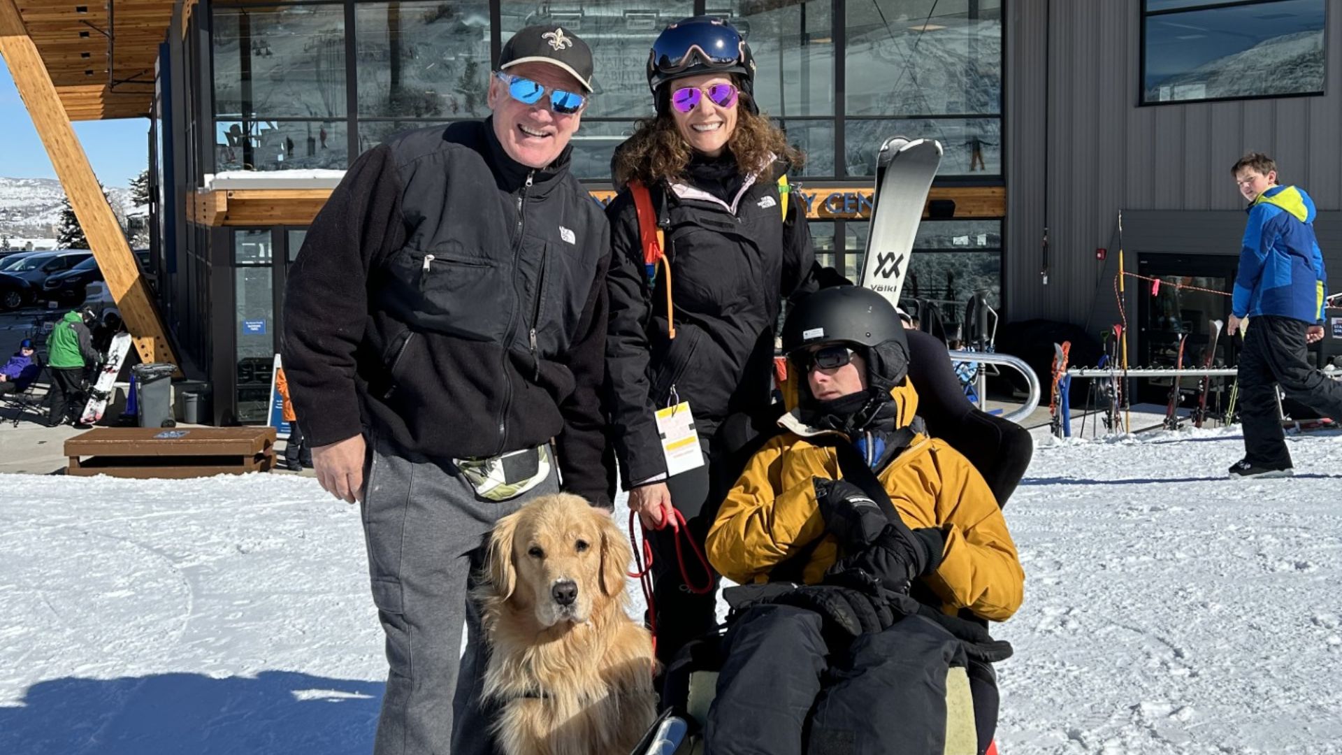 Three people, one seated in adaptive ski equipment, pose outdoors in the snow with ski gear and a golden retriever beside them—capturing a moment from Jen's Advocacy story. A modern building and other skiers are visible in the background.