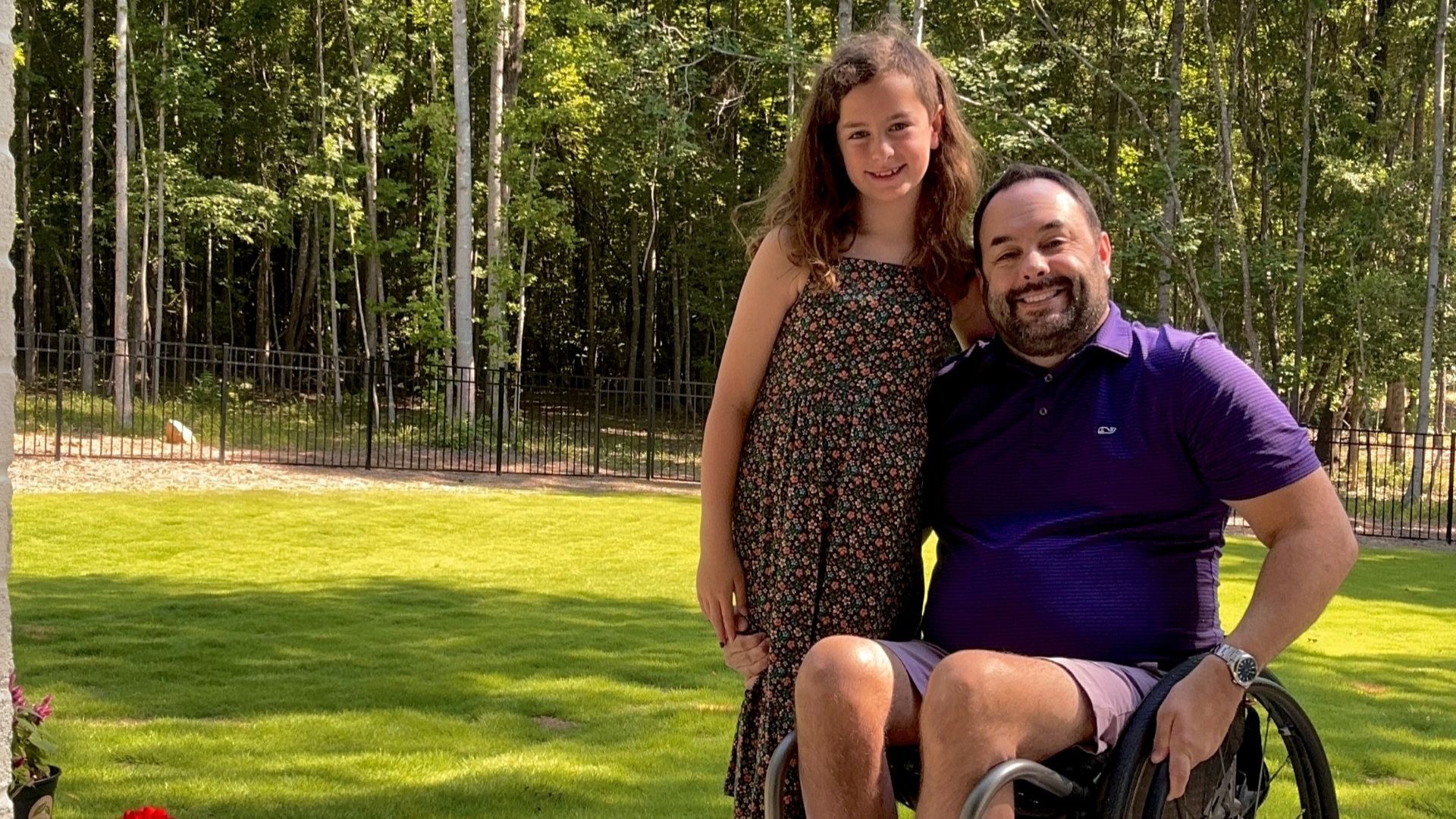 Justin’s advocacy story shines as he smiles in his wheelchair beside a young girl, both enjoying a sunny day outdoors with green grass and trees in the background.