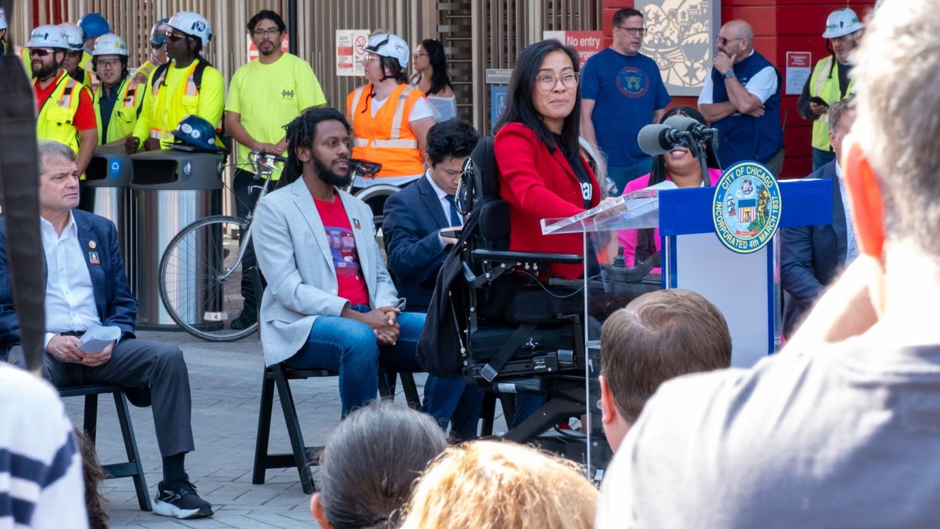 Michele, known for her strong advocacy, speaks at a podium in a red blazer during an outdoor event, surrounded by seated people and construction workers in safety vests and helmets. A crowd watches from the foreground.