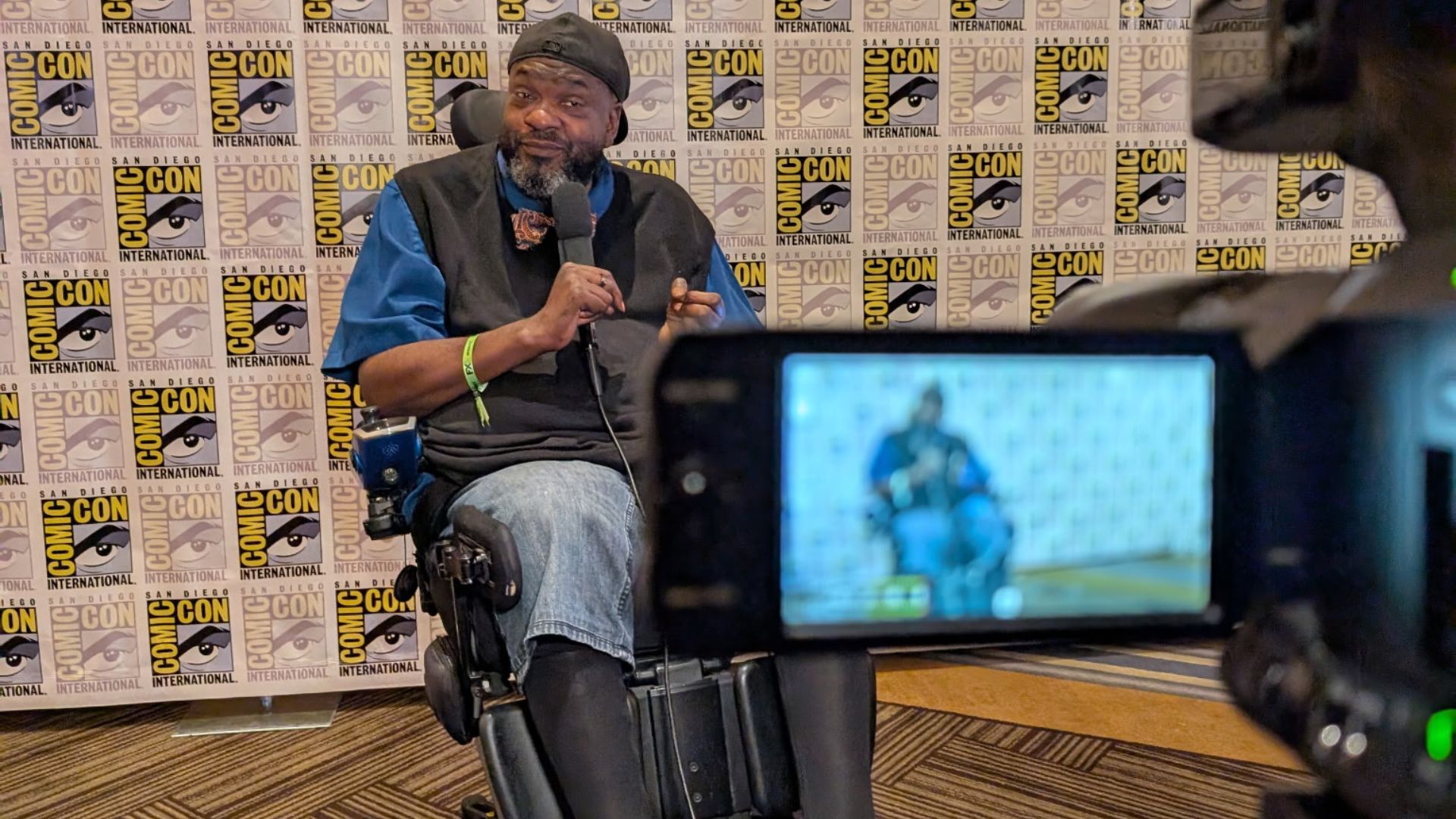 A man in a wheelchair speaks into a microphone at Comic-Con International, with a Comic-Con logo backdrop behind him. Paul's Advocacy shines as a camera in the foreground records the scene, showing his image on its screen.