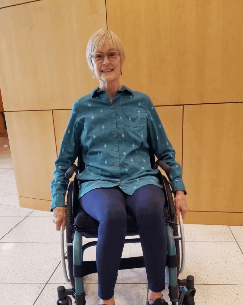 An older woman with short blonde hair, glasses, and a teal button-up shirt sits smiling in a wheelchair in front of a light wood-paneled wall.