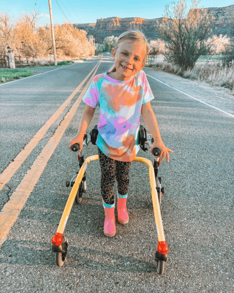 A smiling young girl stands on a road using a yellow walker. She wears a colorful tie-dye shirt, patterned leggings, and pink boots. Trees and rocky hills are visible in the background under a clear sky.