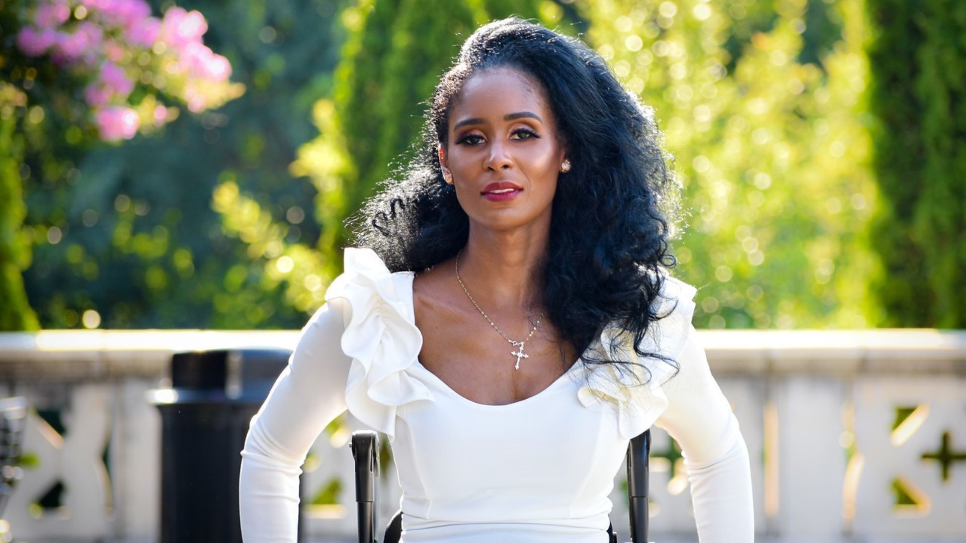 A woman named Tae with long curly black hair, wearing a white ruffled top and a cross necklace, sits outdoors in a sunlit garden, surrounded by greenery and flowers, ready to share her story of advocacy.