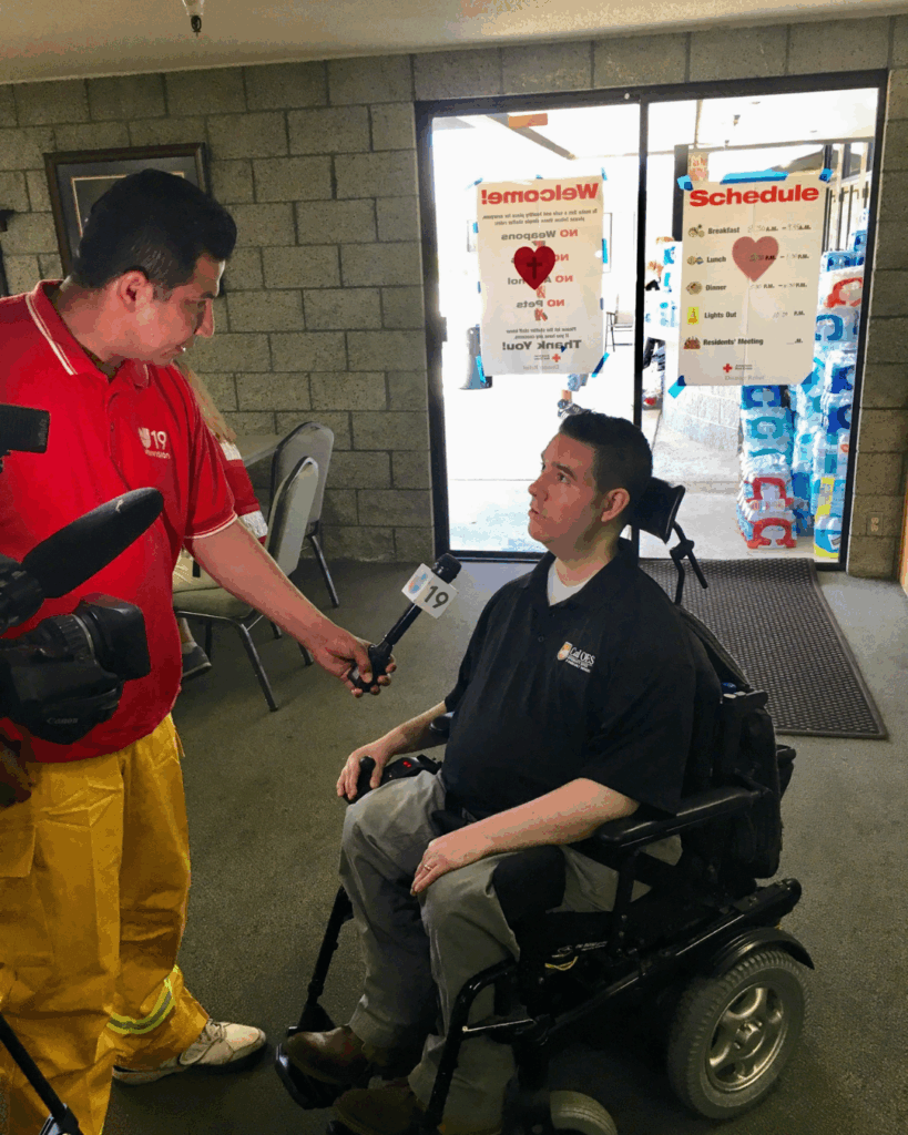 A man in a wheelchair shares Vance's Advocacy Story while being interviewed by a news reporter indoors, with two posters on the glass doors behind him and cases of bottled water stacked nearby.