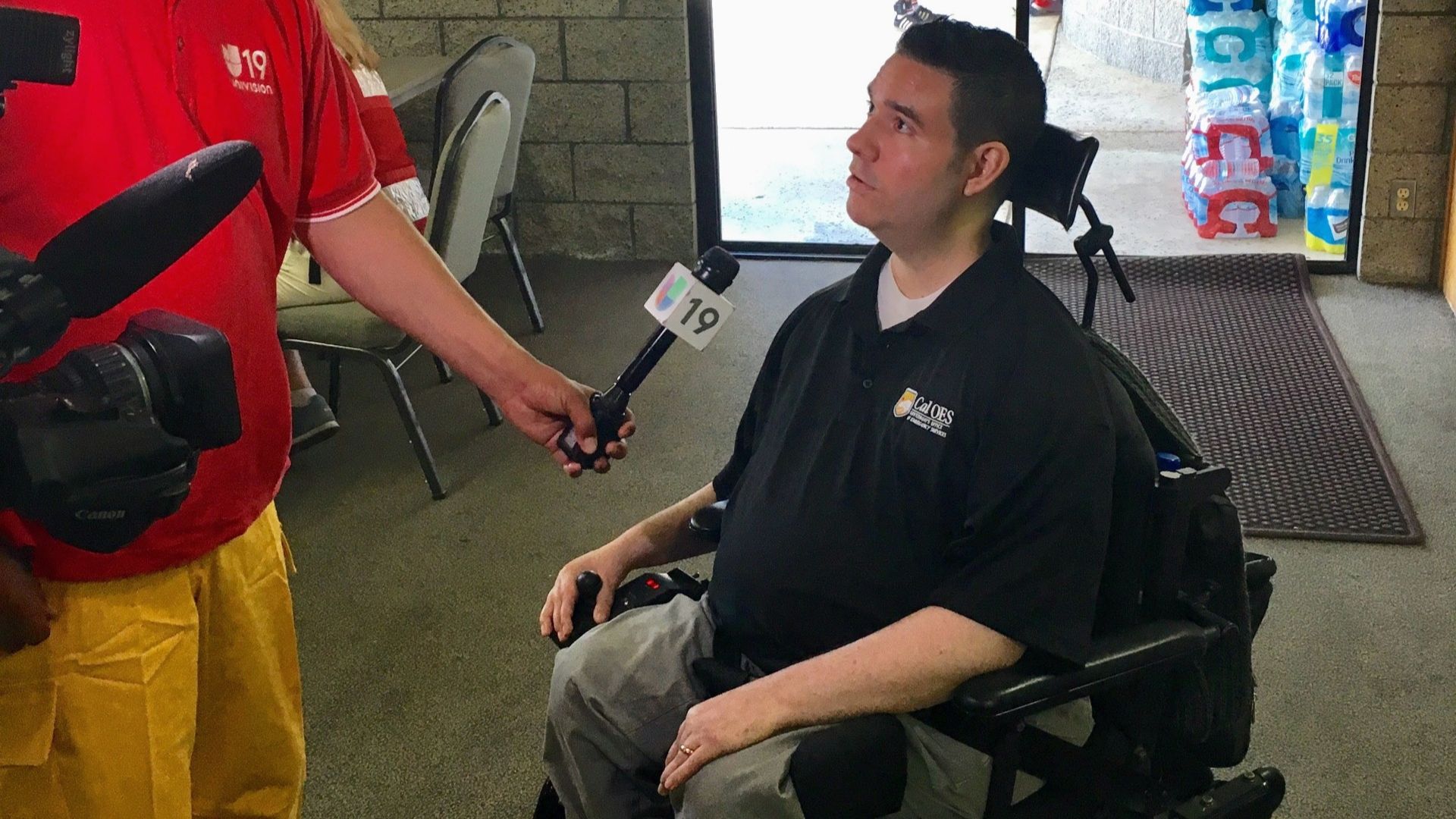 A man named Vance in a wheelchair is being interviewed by a reporter holding a microphone labeled 19. Wearing a black polo shirt, Vance shares his advocacy story as bottled water is stacked in the background.