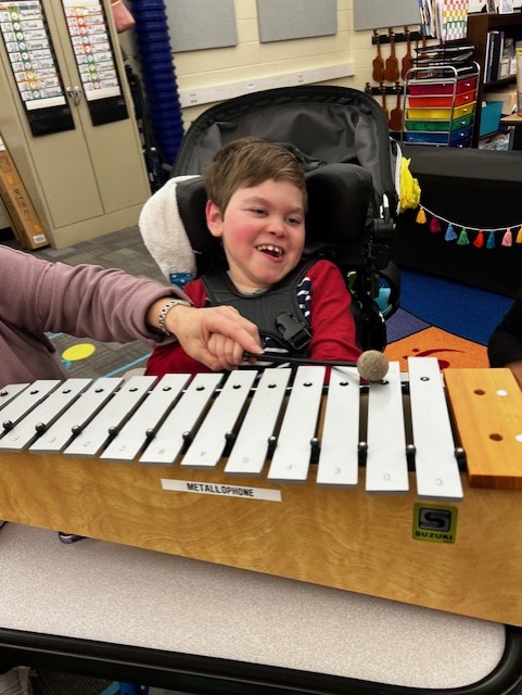 In a colorful classroom, Jake's Story unfolds as a smiling boy in a wheelchair plays a metallophone, his hand gently guided by an adult.